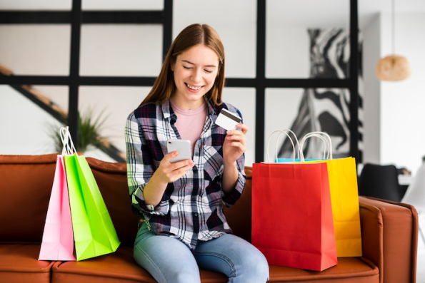 A young woman holding her credit card and smartphone, surrounded by shopping bags, engaging in online shopping