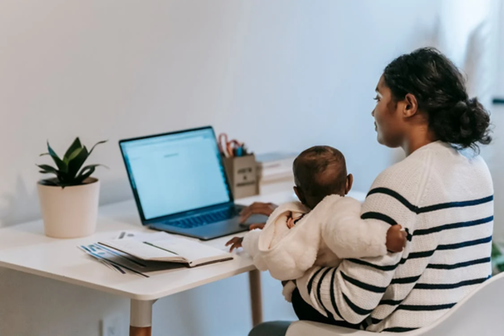 A mother holding her baby while working on a laptop at a white desk.