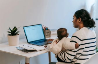 A mother holding her baby while working on a laptop at a white desk.